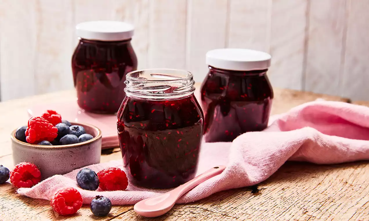 Három üveg sötétbordó áfonya-málna dzsem szálas textúrával egy faasztalon, friss bogyósokkal és puha rózsaszín kendővel körülvéve