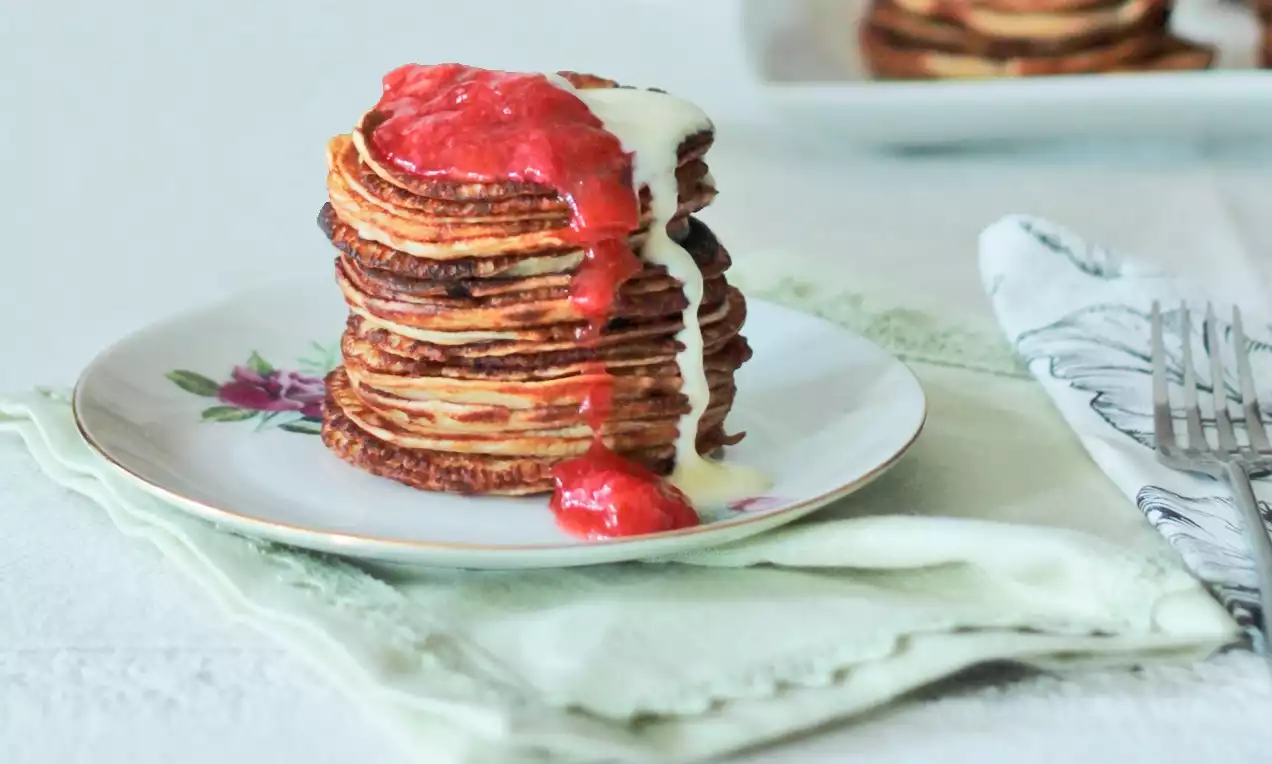 Kullanruskea lettupino, johon valuu mansikanpunainen kastike ja kerma, aseteltuna kukkakoristeiselle lautaselle vaalean liinan päälle