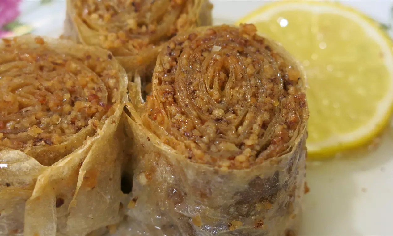 Baklava u obliku čvrsto urolanih rolnica zlatnosmeđih tonova, sa zrnastom teksturom, oslonjena uz krišku svežeg limuna na tanjiru
