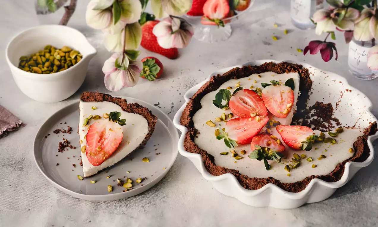 Maskarpones deserts ar tumšu, drupenu cepumu pamatni, krēmīgu baltu pildījumu, dekorētu ar spilgti sarkanām zemenēm un zaļiem pistāciju gabaliņiem