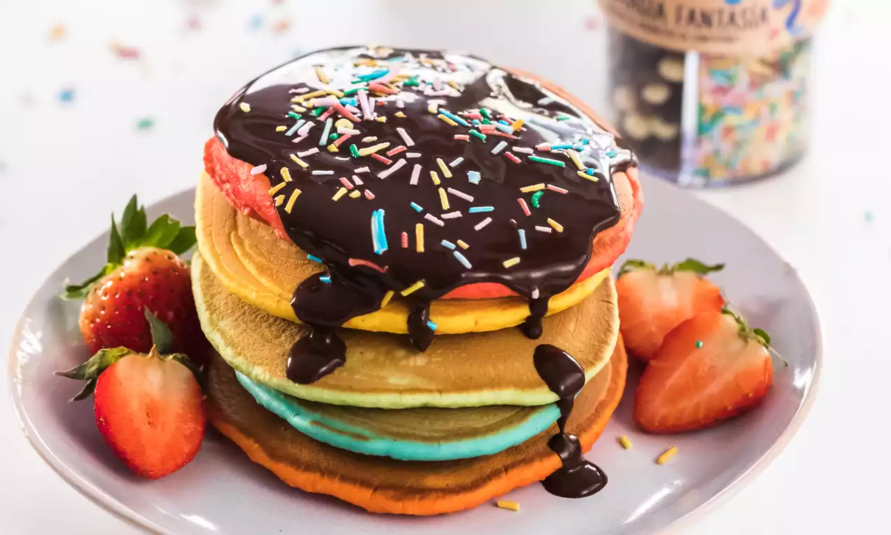 Tortitas de colores vibrantes en capas verticales, bañadas con salsa de chocolate brillante y decoradas con chispas multicolor junto a fresas partidas