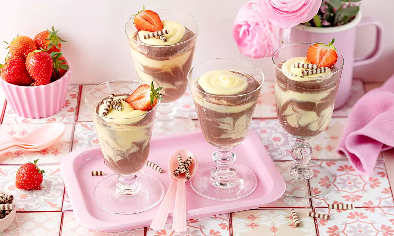 Schoko-Vanille-Dessert im Glas mit marmorierter Creme, frischen Erdbeeren und Schokoröllchen, arrangiert auf rosa Tablett mit dekorativen Details