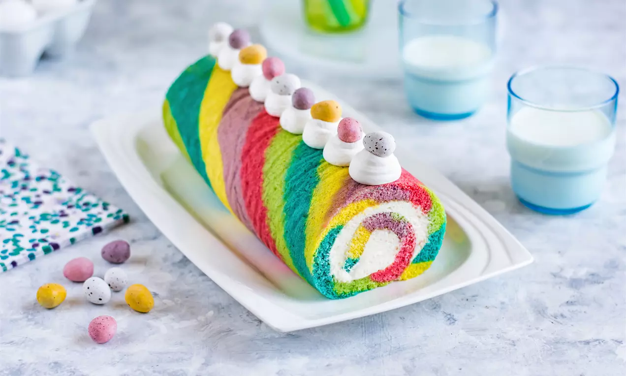 Руло с разноцветна шарка в зелено, жълто, розово и синьо, декорирано с бели розички от крем и захарни яйчица върху бяла чиния