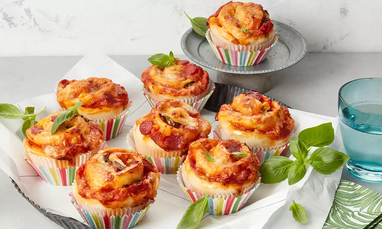 Pizza muffins aurii cu topping de mozzarella topită, felii de ceapă și sos roșu, aranjate pe hârtie albă, decorată cu frunze de busuioc verde