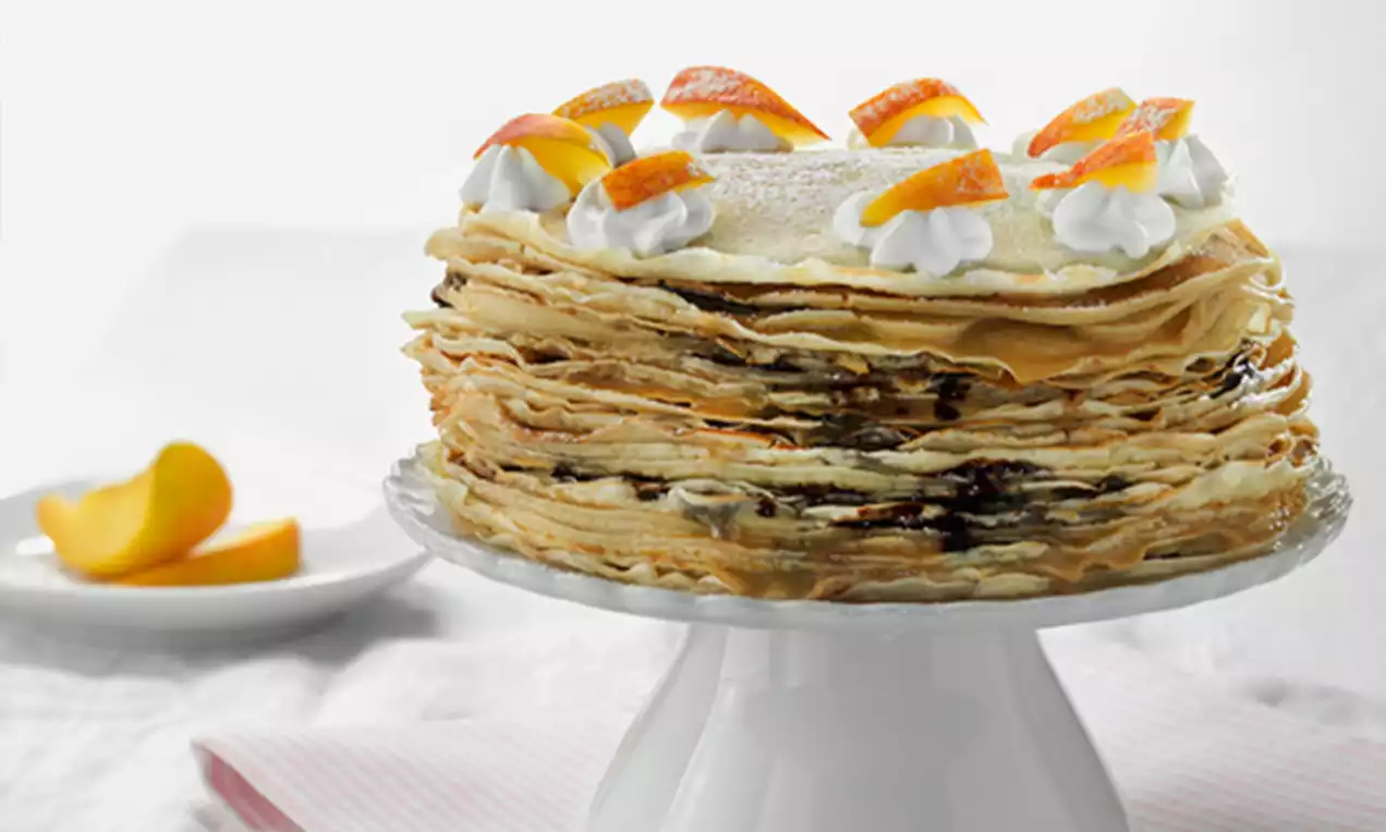 Tarta dorada de capas finas de crepes rellenas de crema, decorada con rosetones de nata y rodajas de naranja sobre un plato blanco
