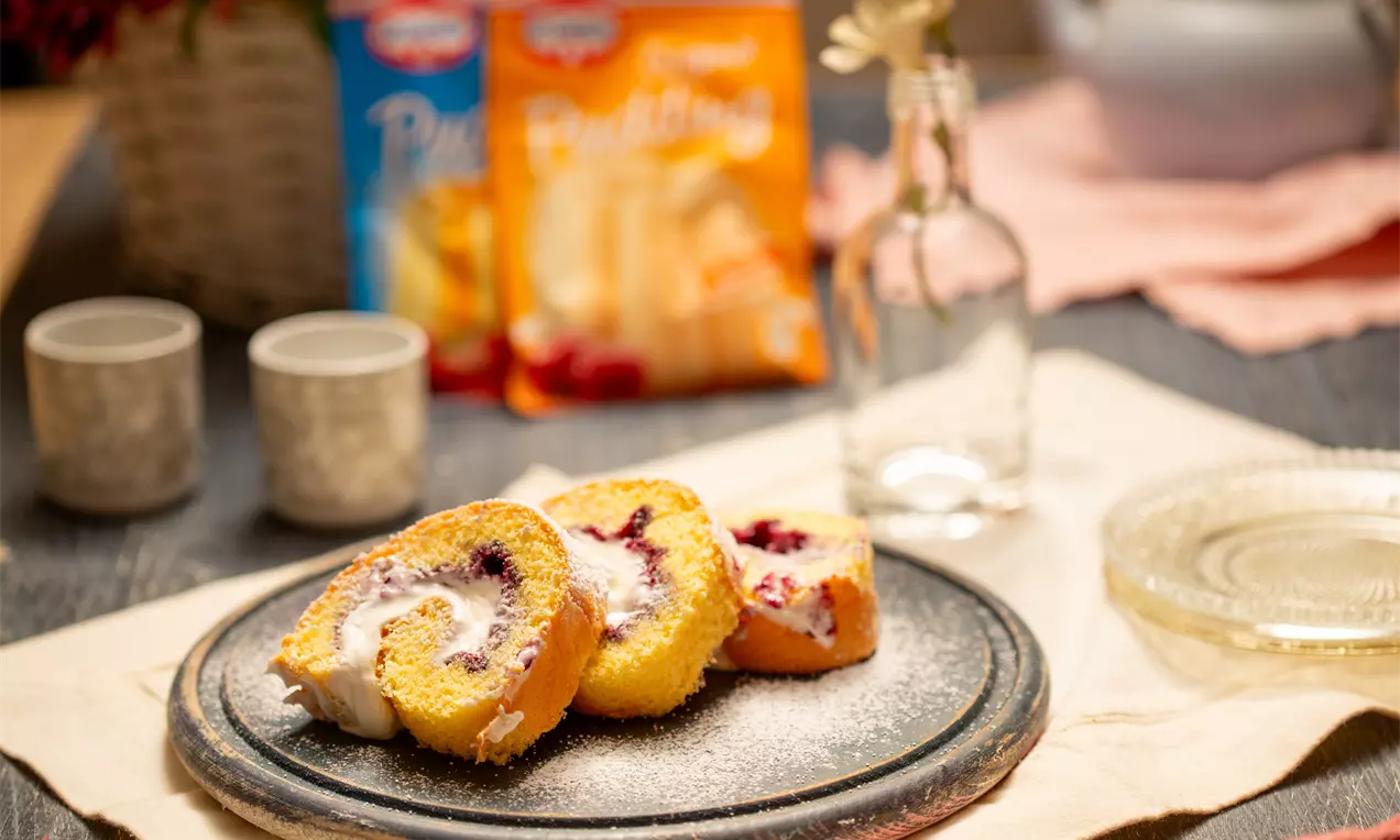 Златисто руло с лека пухкава текстура и завит сметанов крем с яркочервени плодови акценти, нарязано и сервирано върху тъмна чиния