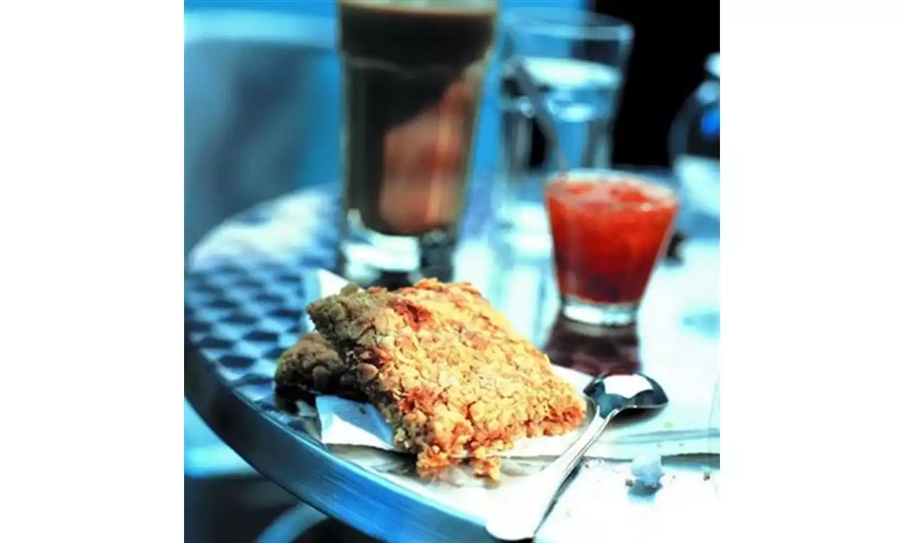 To gyllenbrune havrecrunchies med smuldrete tekstur ligger på hvitt papir ved siden av en liten skje på et metallbord med drikke i bakgrunnen