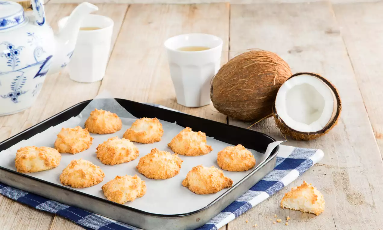 Goudbruine, ronde kokoskoekjes met een licht gebakken textuur op bakpapier in een zwart bakblik, naast een halve kokosnoot en witte kopjes