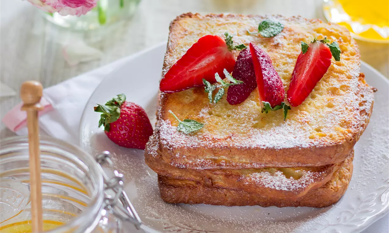 Пухкави златисти филии френч тост, подредени една върху друга, гарнирани със сочни парчета ягоди и листенца мента, поръсени с пудра захар