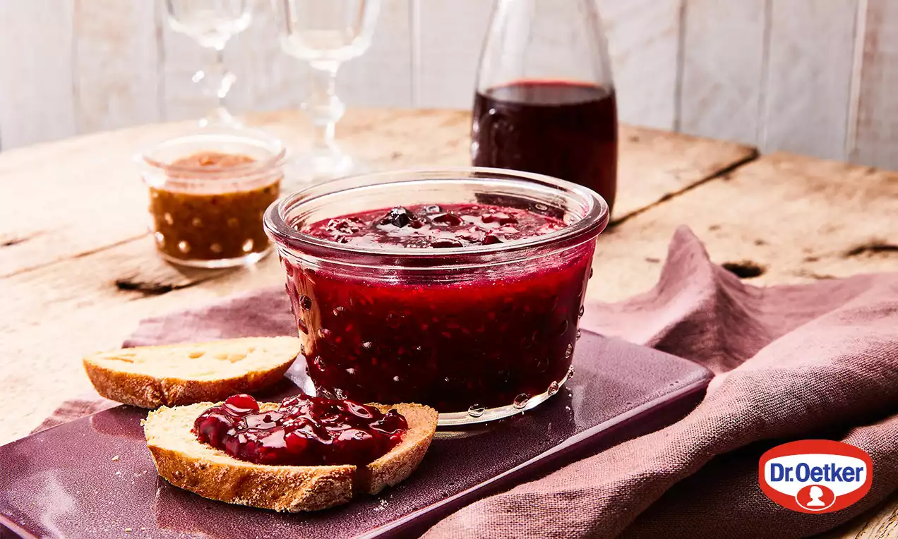 Hustý lesklý catni tmavě červené barvy s drobnými kousky ovoce v průhledné sklenici na fialovém podnosu vedle světlého chleba