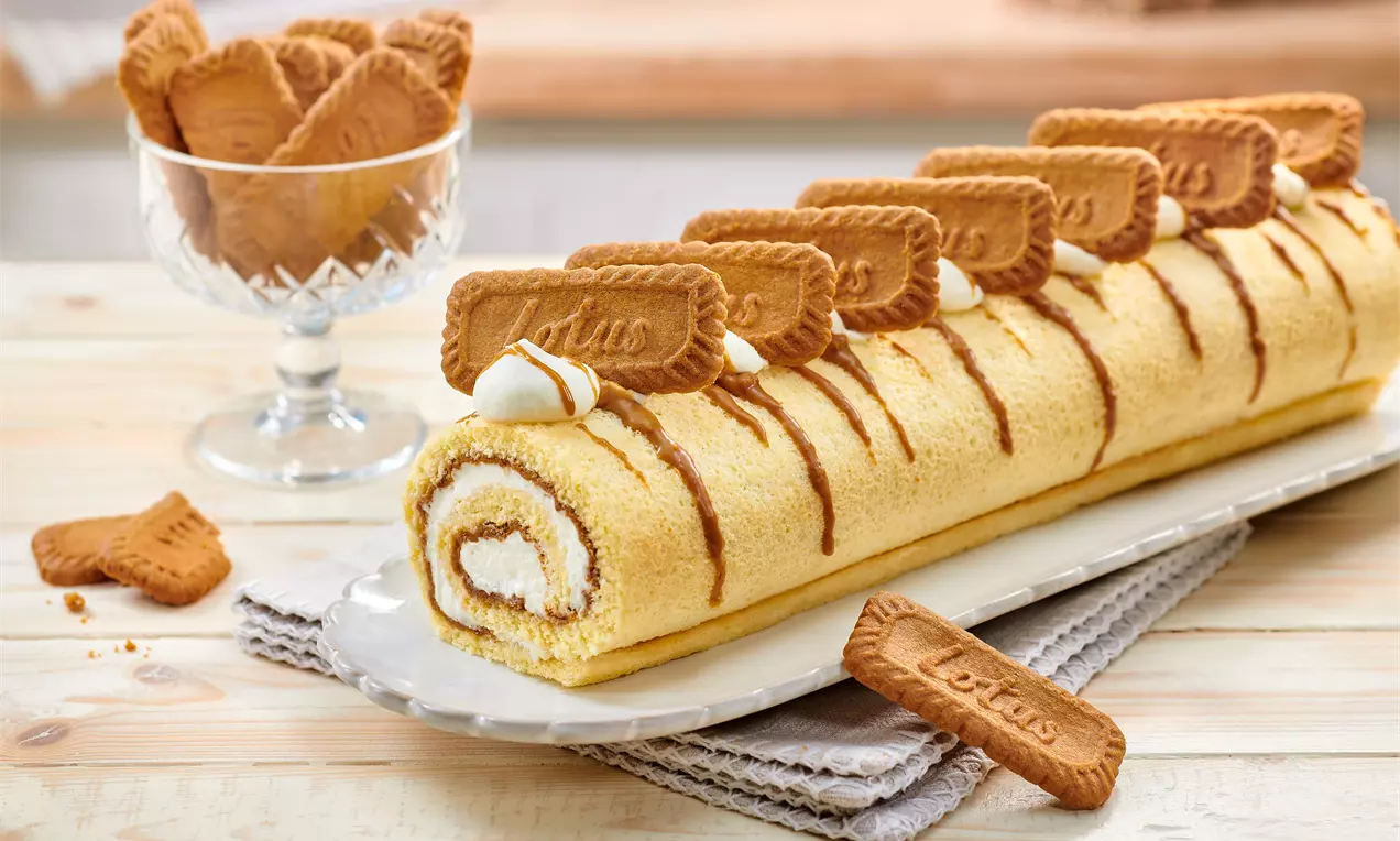 Goldgelbe Biskuitrolle mit weißer Sahnefüllung, dekoriert mit Karamellstreifen und Lotus-Keksen, auf einer weißen Platte angerichtet