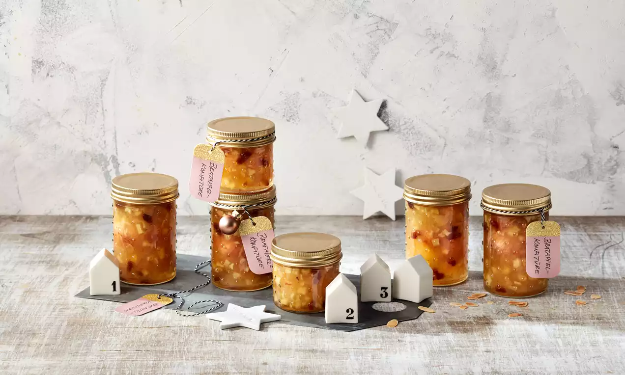 Bratapfel-Konfitüre in golden verschraubten Gläsern mit sichtbaren Fruchtstückchen, auf Holzplatte dekorativ arrangiert mit Sternen und Zahlenhäusern