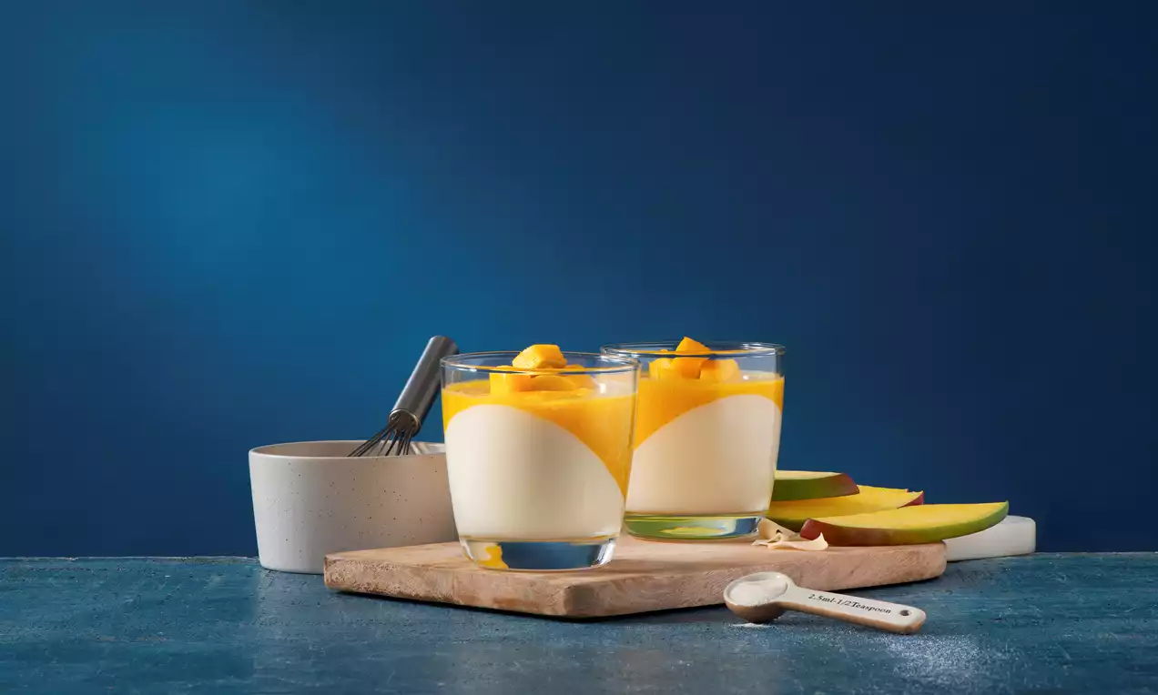 Two glass cups of creamy white panna cotta curved with golden mango puree and diced mango on a wooden board with a spoon and sliced fresh mango