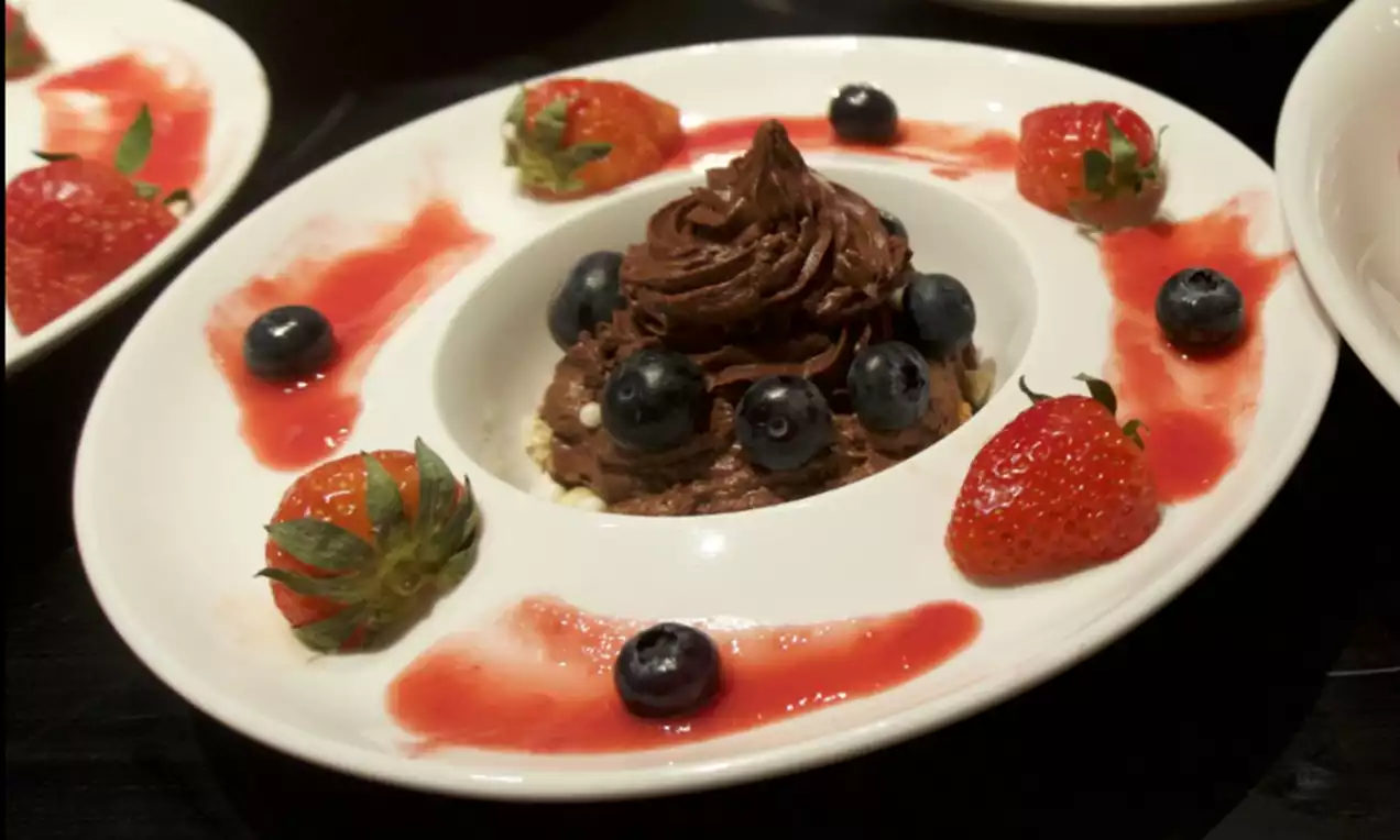 Chocolademousse in een torentje met glanzend bruine krullen, omringd door blauwe bessen, aardbeien en rode saus op een wit bord