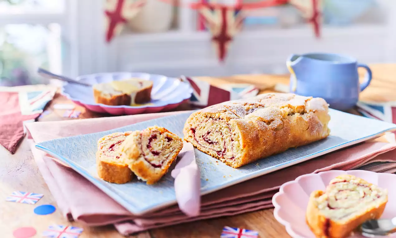 Golden jam roly poly with swirls of glossy red jam on a rectangular pale blue plate, dusted with sugar, surrounded by Union Jack decor