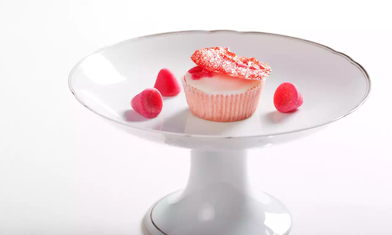 Cupcake met lichtroze glazuur en suikerdecoratie, omgeven door drie kegelvormige roze cuberdons op een wit sierlijk taartplateau