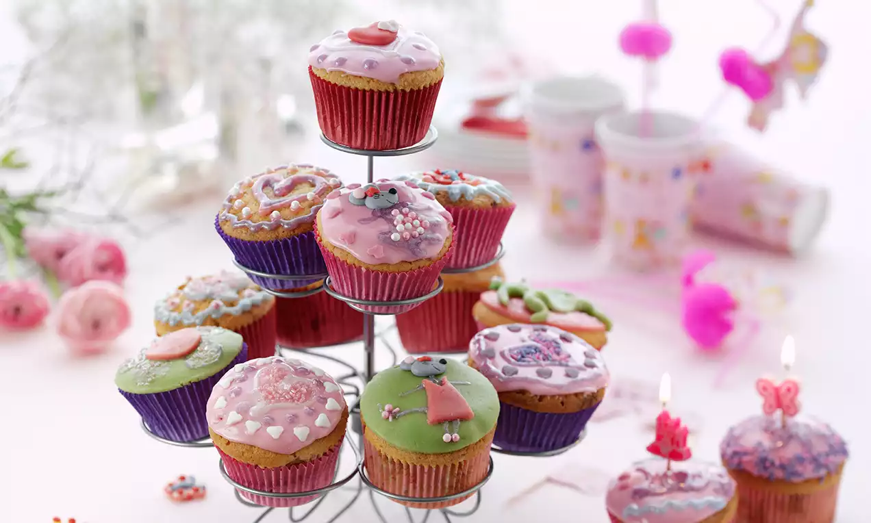 Muffins mit bunter Glasur in Rosa, Grün und Lila, verziert mit Zuckerstreuseln und figürlichem Dekor auf silbernem Ständer präsentiert