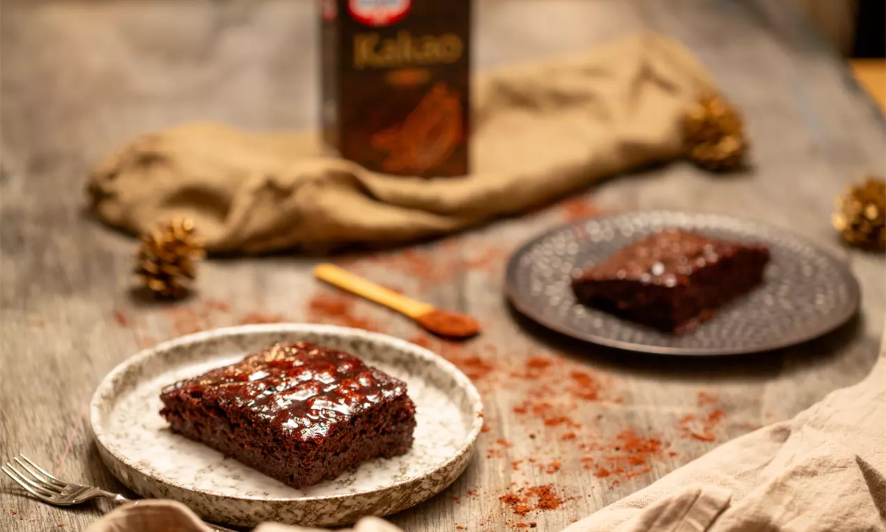 Квадратно парче тъмен шоколадов фъч с плътна текстура и лъскава глазура, поднесено върху светла чиния на дървена маса