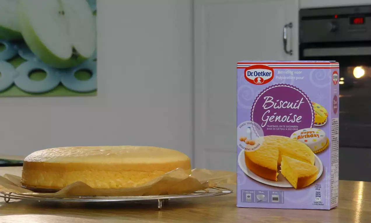 Lichtbruine, luchtige biscuitcake op bakpapier en rooster, naast een paarse verpakking met afbeelding van een vergelijkbare cake in een keuken