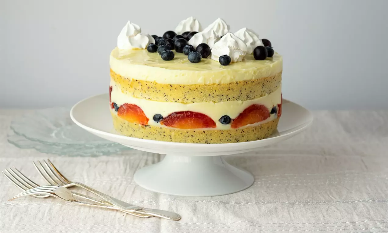 Слоеста лимонова торта с маково семе, кремообразен пълнеж, свежи плодове и целувки, поднесена върху бяла порцеланова поставка