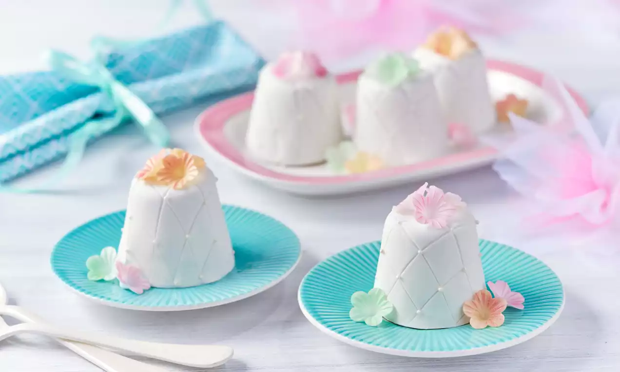 Mini torciki w białej polewie z subtelnym wzorem kratki, ozdobione kolorowymi kwiatkami, na pastelowych talerzykach i jasnym tle