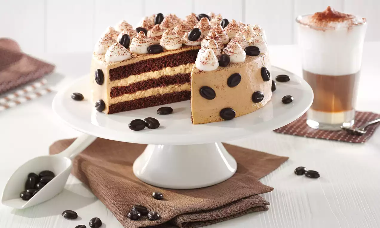 Kleine Latte-Macchiato-Torte mit Schichten aus braunem Teig und heller Creme, dekoriert mit Sahnetupfen, Kakaopulver und dunklen Kaffeebohnen