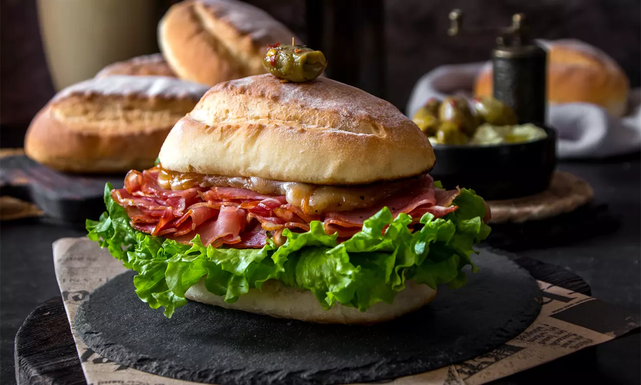 Златистокафява питка с хрупкава кора, пълна със сочни резени месо, пресни зелени листа салата и кисела краставичка върху черна поставка