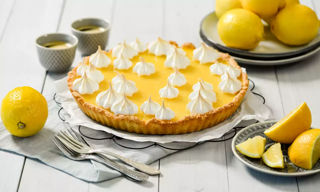 Zitronentarte mit gelber Creme und weißer Baiserhaube auf Backpapier, umgeben von frischen Zitronen und Gabeln auf einem weißen Holztisch