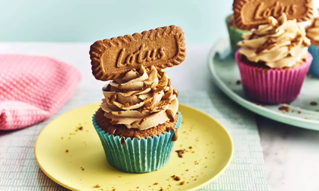 Golden-brown cupcakes in blue and pink wrappers topped with swirls of caramel-coloured cream and a Lotus biscuit on pastel plates