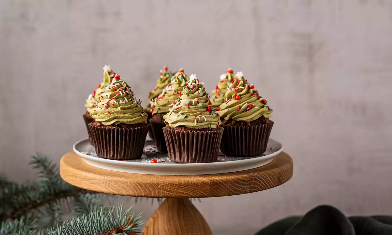 Šokoladiniai keksiukai su banguotu žaliu kremu, primenančiu eglutes, papuošti spalvotais pabarstukais, medinėje lėkštėje ant kojelės