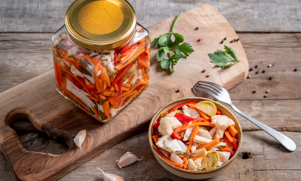 Цветна салата от бели карфиолови розички, оранжени резени моркови и червени парчета чушка, подредени в купа до буркан върху дървена дъска