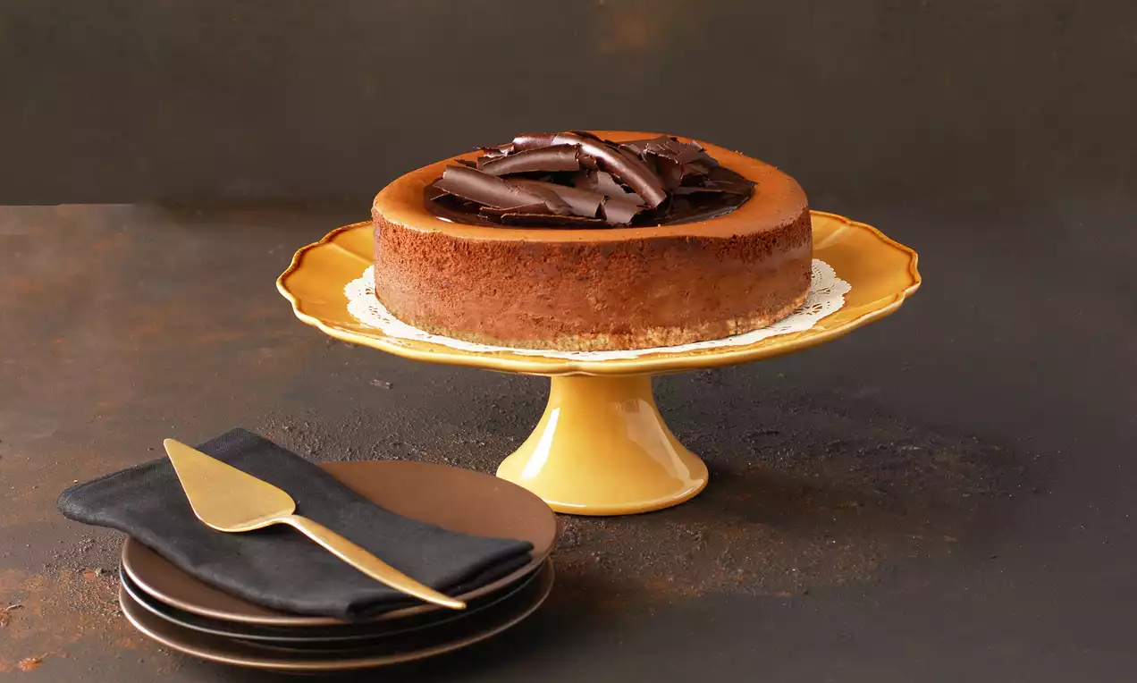 Kahverengi tabanı ve yoğun dokusuyla göz alıcı çikolatalı bademli cheesecake, sarı ayaklı bir tabakta çikolata parçalarıyla süslenmiş