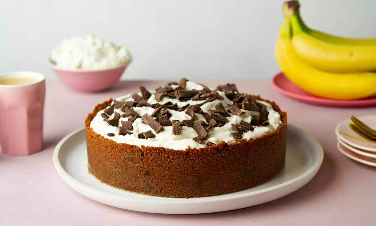 Banoffee-taart met bruine krokante koekbodem, luchtige witte slagroomlaag en donkere chocoladestukjes op een licht bord met roze en gele decoratie