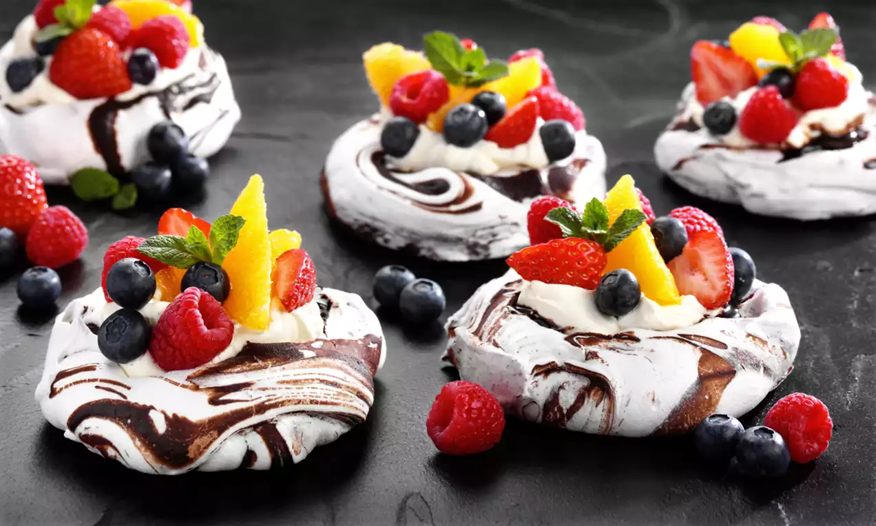 Chocolate marbled meringue nests with glossy swirls, topped with whipped cream, vibrant berries, orange segments and mint sprigs on a dark surface