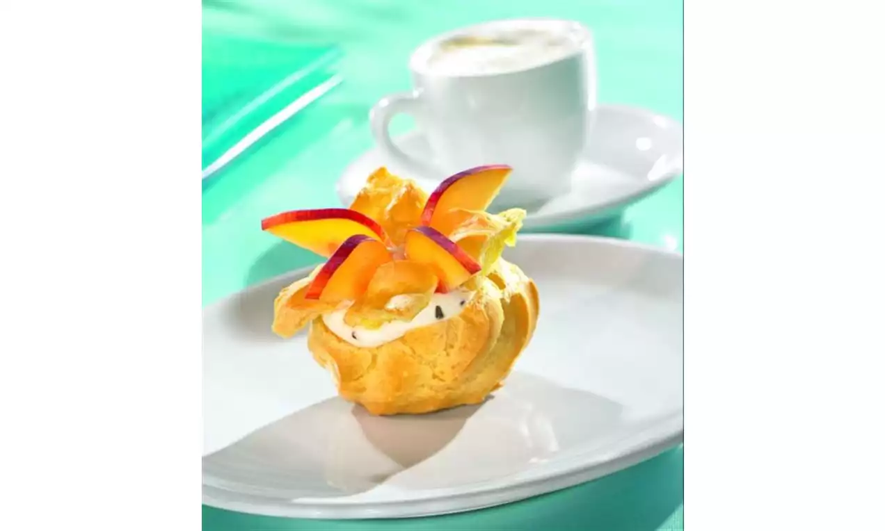 Chou doré garni de crème blanche, décoré de tranches de pêche orange et rouge, posé sur une assiette blanche avec une tasse en arrière-plan