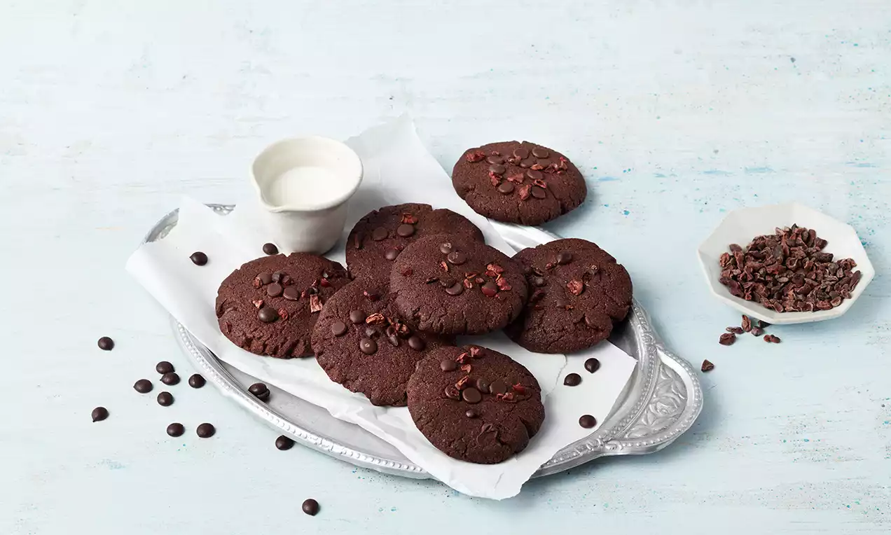 Dunkelbraune, runde Schokokekse mit matter Oberfläche und Schokodrops auf weißem Papier, arrangiert auf einem silbernen Tablett mit Soßenbecher