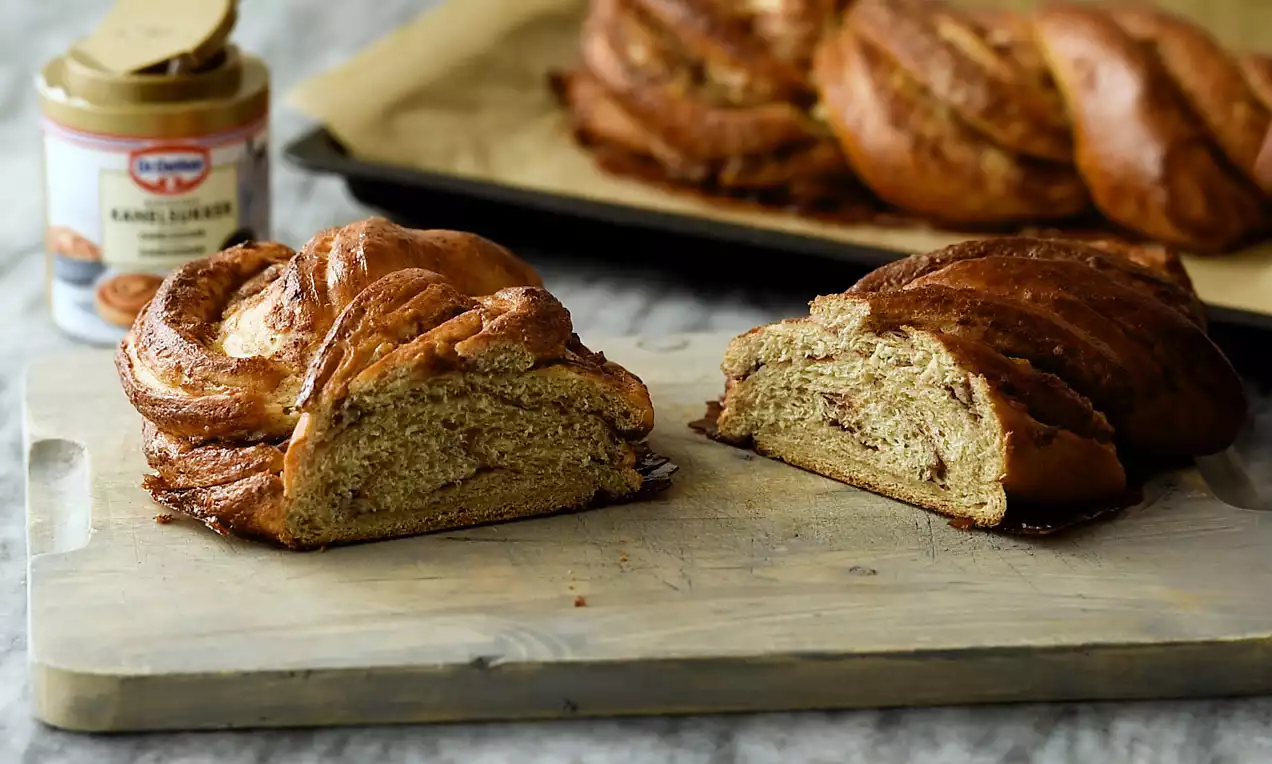 Gyldenbrunt flettet kanelbrød med luftig krumme og kanelswirls, skåret i halve på et lyst træbræt med et brød og kanelsukker bagved