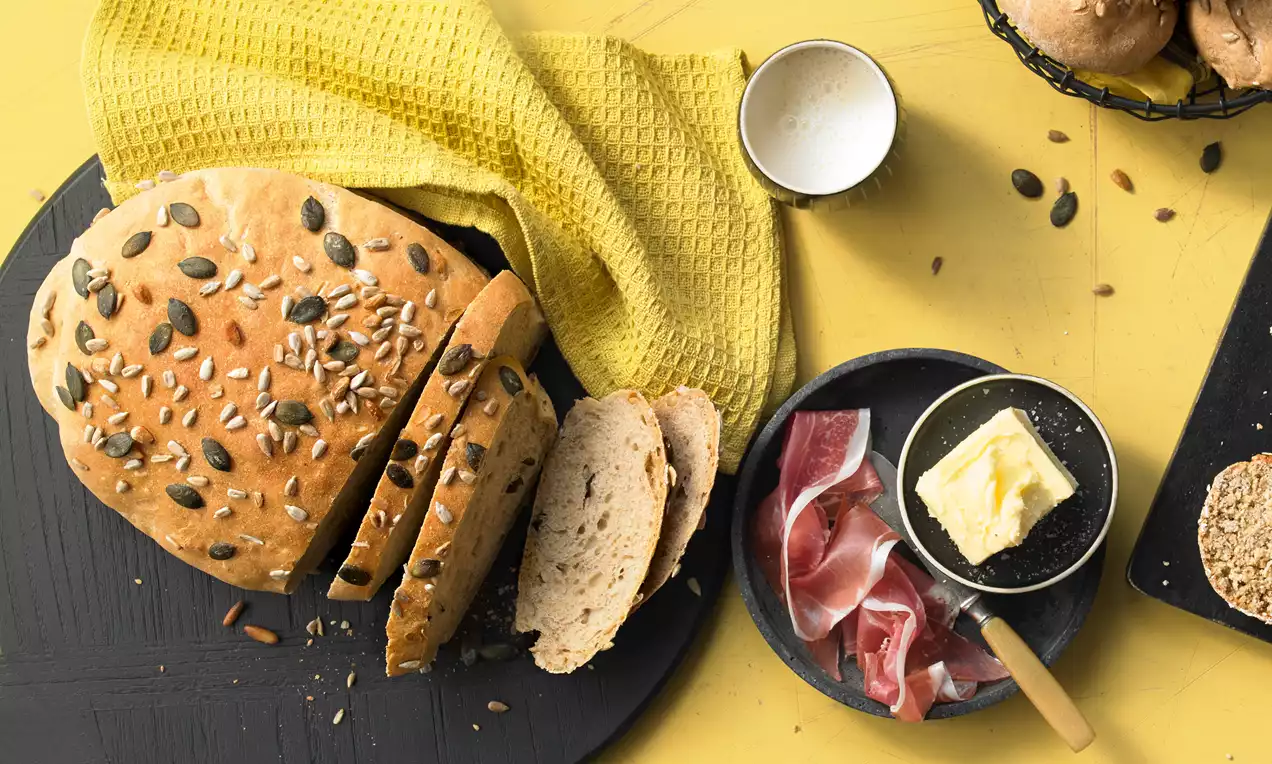 Dinkelbrot mit goldbrauner Kruste und Körnern bestreut, in Scheiben geschnitten auf dunklem Holzbrett mit Butter, Schinken und gelbem Tuch drapiert