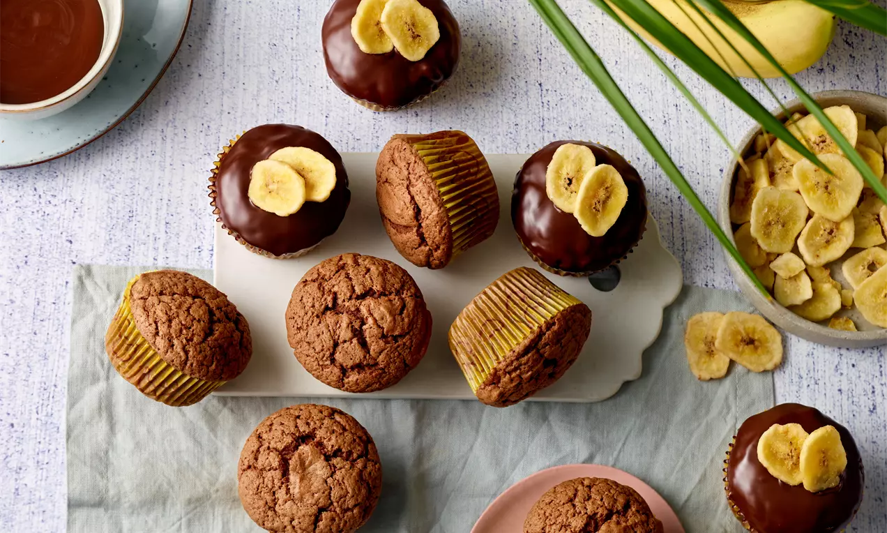 Muffins maro-aurii pufoși, unii glazurați cu ciocolată lucioasă și decorați cu felii de banană, așezați pe platouri albe și roz