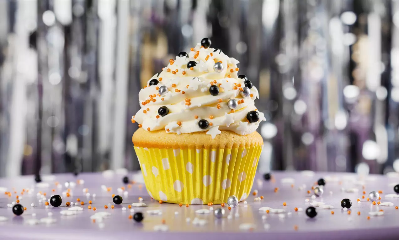 Cupcake met een luchtige witte toef versierd met zwarte, zilveren en oranje pareltjes in een geel gestipt vormpje op een paarse ondergrond