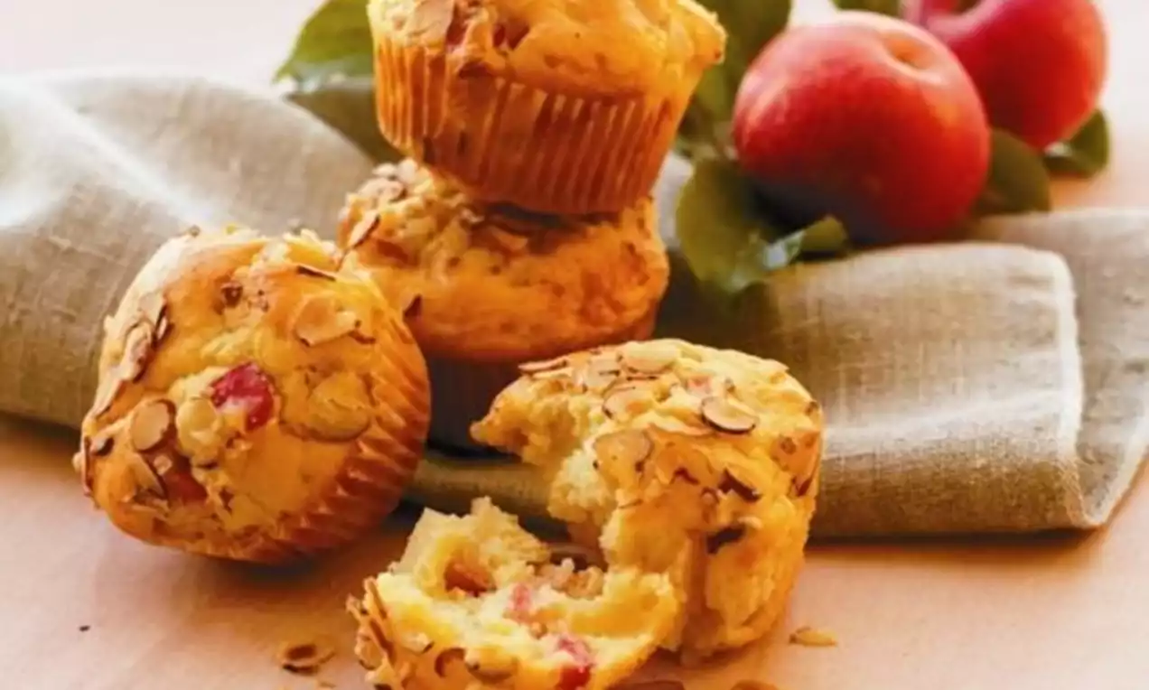Eplemuffins gyllenbrune med skiver av mandler, en muffins delt i to på beige duk med røde epler og grønne blader i bakgrunnen