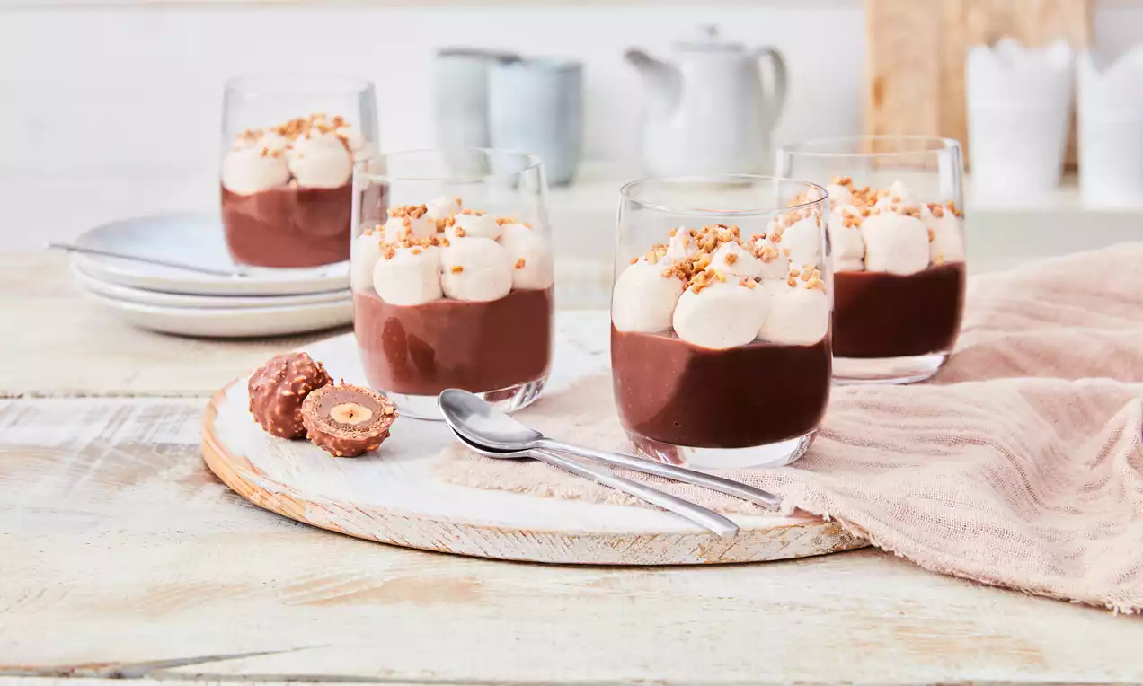 Pudim cremoso de chocolate escuro em copos transparentes decorado com camadas de chantilly branco e avelãs moídas sobre mesa de madeira clara