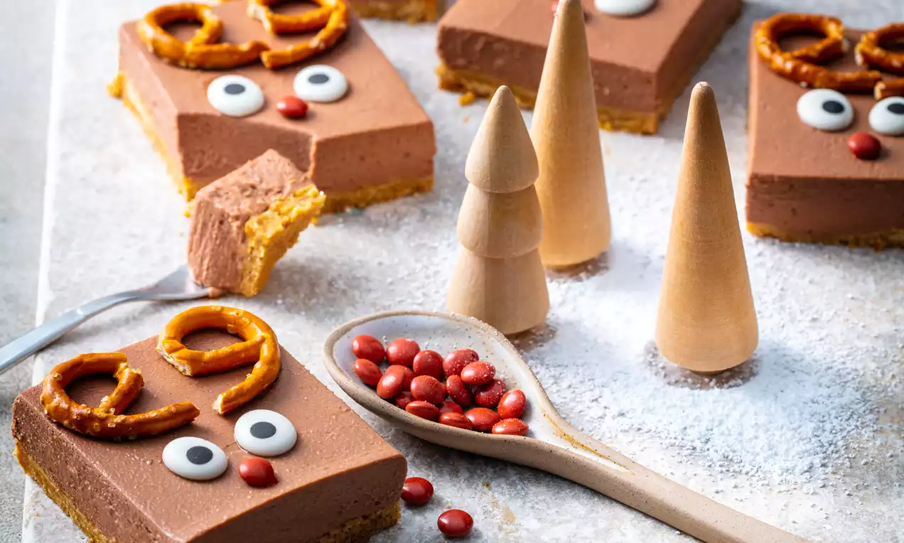Carrés crémeux au chocolat sur base biscuitée, décorés de bretzels dorés, yeux sucrés et nez rouge, avec cônes et billes rouges sur un plateau