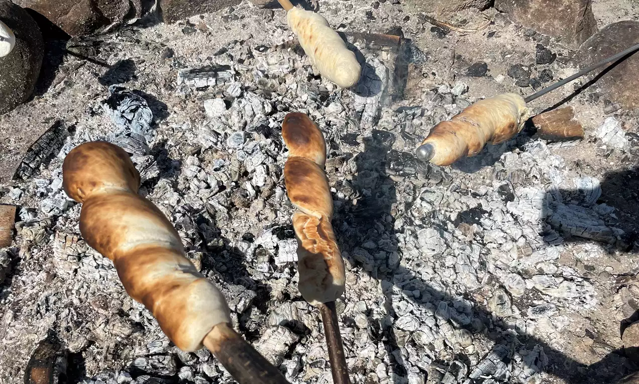 Snobrød brunsprø og gyldne på trekkerter, vridd rundt pinnene over glødende grå aske og kull med steiner i bakgrunnen
