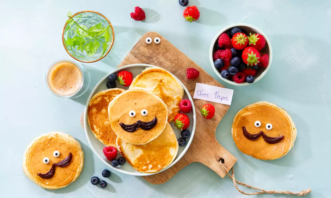 Goudbruine pannenkoekjes met chocoladesnorren en oogjes op een houten plank, omringd door rood en blauw fruit, met een kaartje voor papa