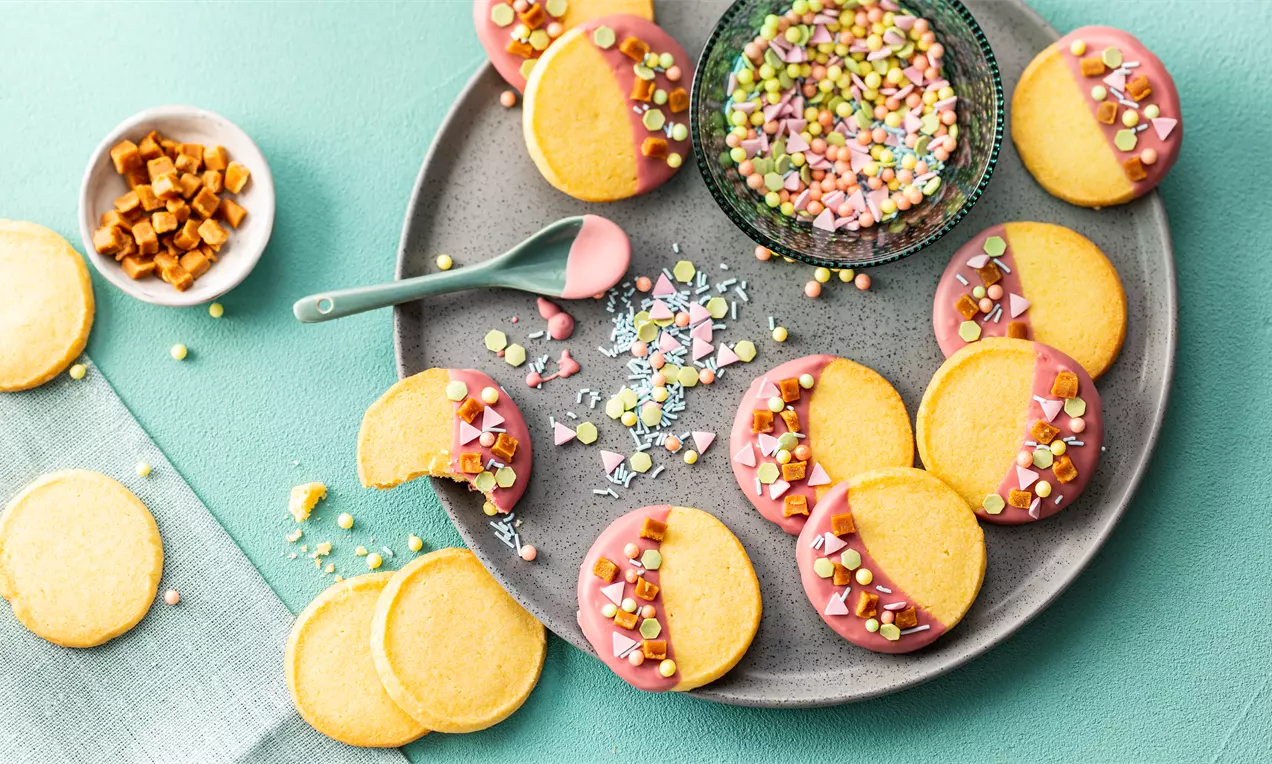 Кръгли ванилови бисквити с гладка златиста повърхност, потопени наполовина в розова глазура с разноцветни захарни фигурки върху сива чиния