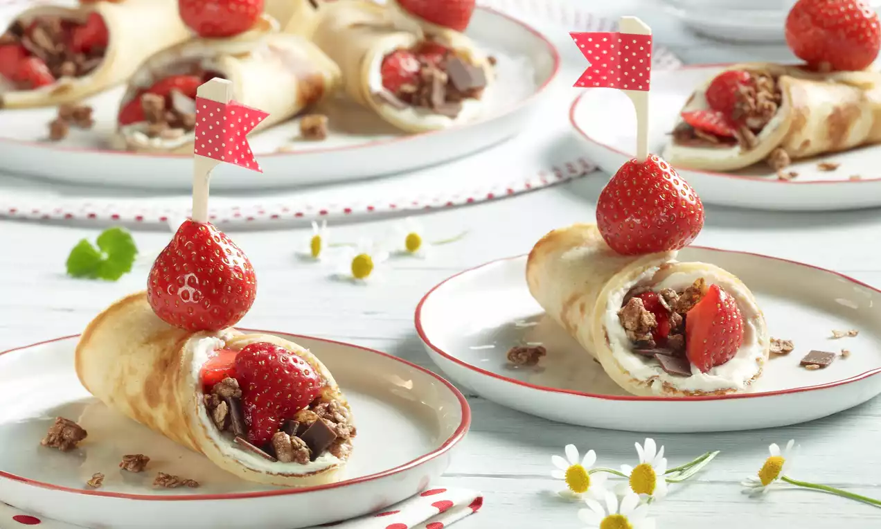Fyldige pannekakerullet med hvit krem, hakkede nøtter og jordbærbiter, toppet med røde jordbær og små flagg, servert på hvite tallerkener