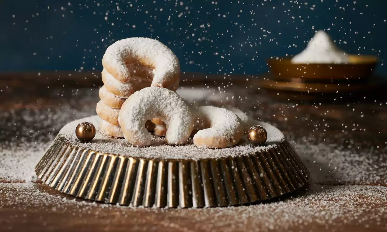 Vanilkové rohlíčky světle zlaté barvy ve tvaru půlměsíce, posypané moučkovým cukrem, umístěné na kovové zlaté míse na dřevěném stole