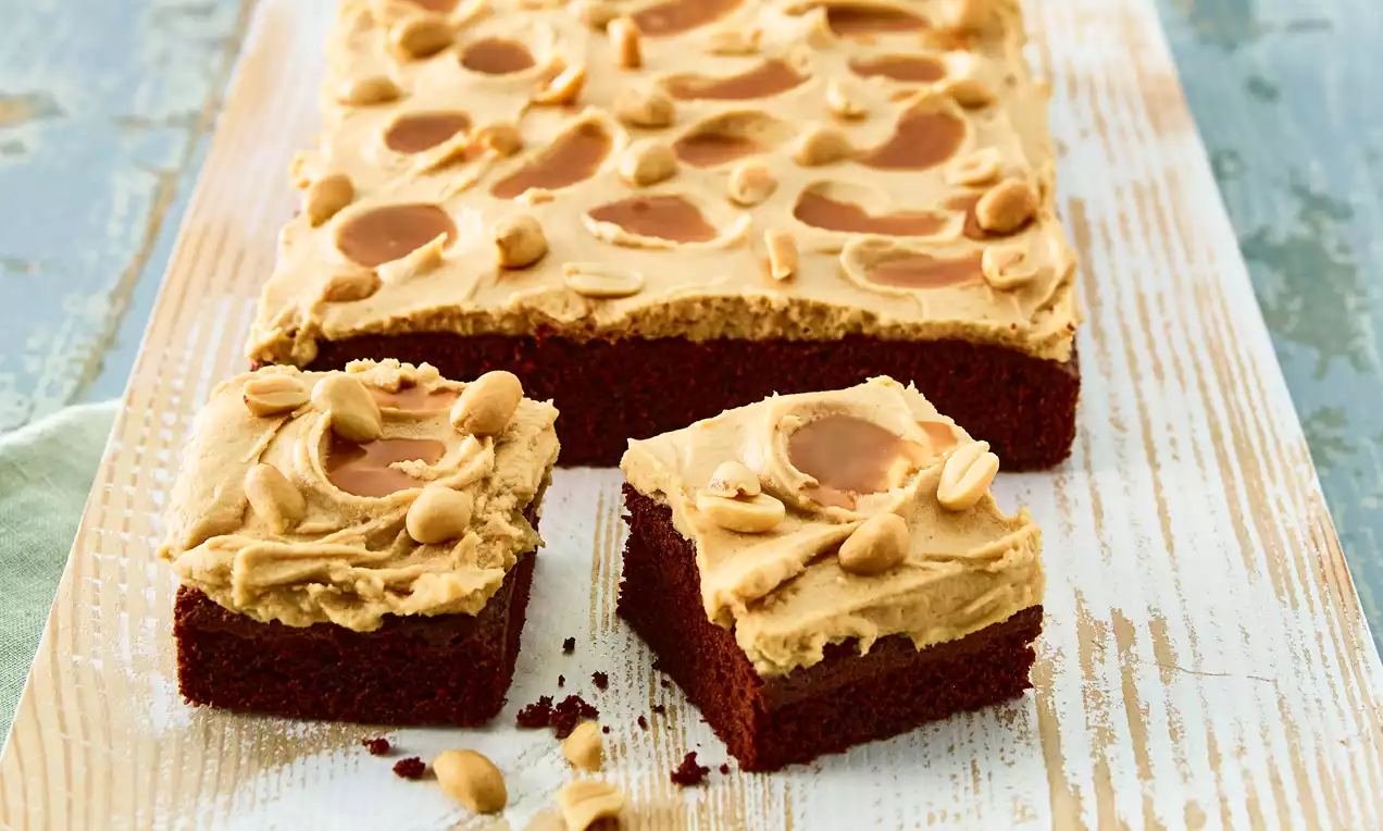 Čokoladni brownies s glatkim premazom od kikiriki-karamel kreme, posuti kikirikijem i garnirani sjajnim karamel mrljama na drvenoj podlozi