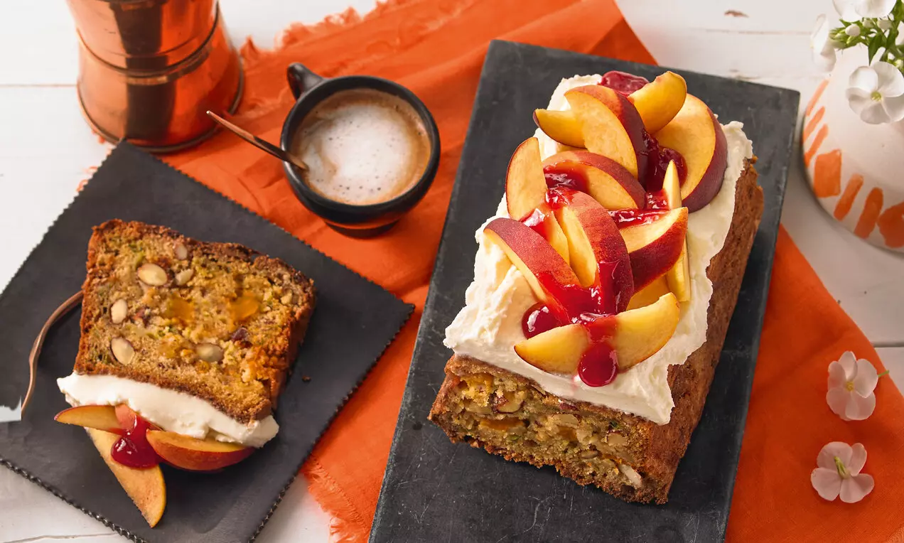 Златисто-кафява плодова торта с тиквички и бадеми, покрита с крем и гарнирана с половинки праскови и гланциран черешов сос върху черна плоча